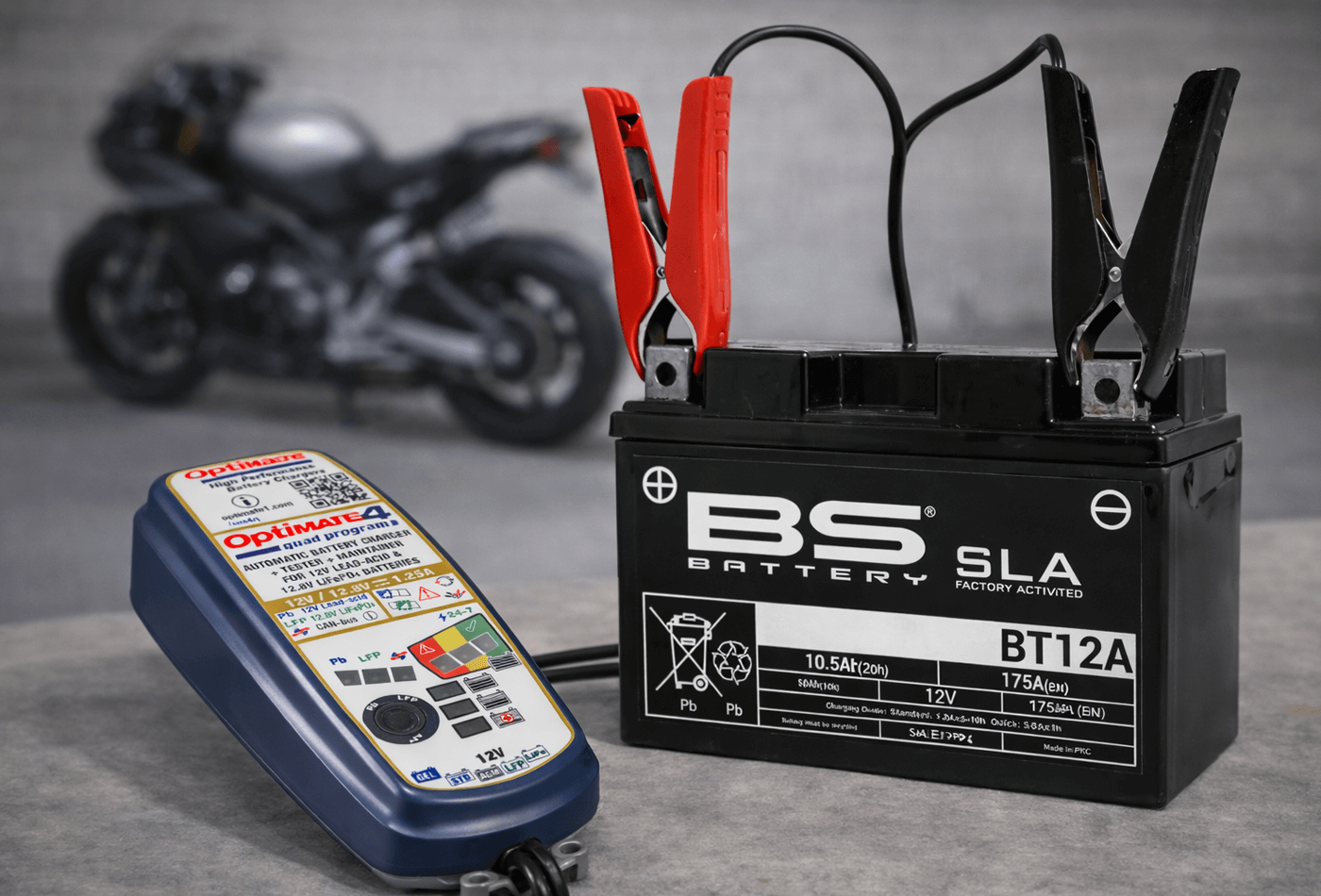 Motocyklová 12 V SLA batéria BS BT12A s inteligentnou nabíjačkou OptiMate 4, v pozadí rozostrená odstavená motorka.
