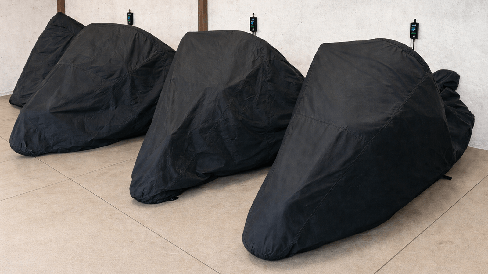 Tri motocykle zakryté čiernymi ochrannými plachtami počas zimného uskladnenia v interiérovej garáži s pripojenými udržiavacími nabíjačkami.